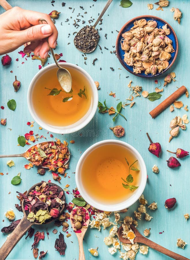 Two cups of healthy herbal tea with mint, cinnamon, dried rose and camomile flowers in spoons and man`s hand holding spoon of honey, blue background, top view. Two cups of healthy herbal tea with mint, cinnamon, dried rose and camomile flowers in spoons and man`s hand holding spoon of honey, blue background, top view