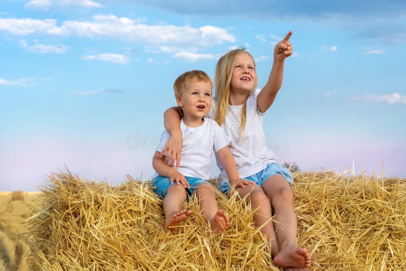 Two cute adorable caucasian siblings enjoy having fun sitting on top over golden hay bale on wheat harvested field near