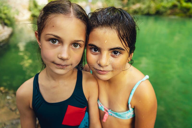 Portrait of a two cute little girls standing near the mountain`s lake, with pleasure spending time in summer camp, happy best friends enjoying summer time together