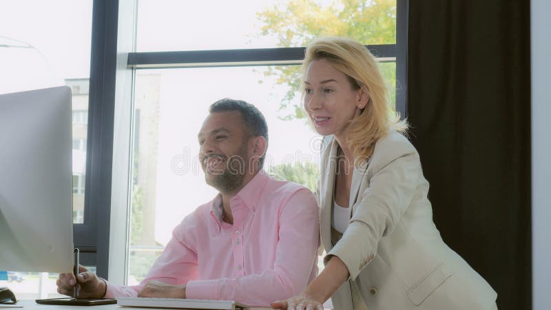 Two designers discussing project. Happy workers looking together on screen computer. Mixed race men drawing on digital tablet. Adult blonde standing near working place and giving advice about logo. Coworkers in modern office with big window talking and smiling