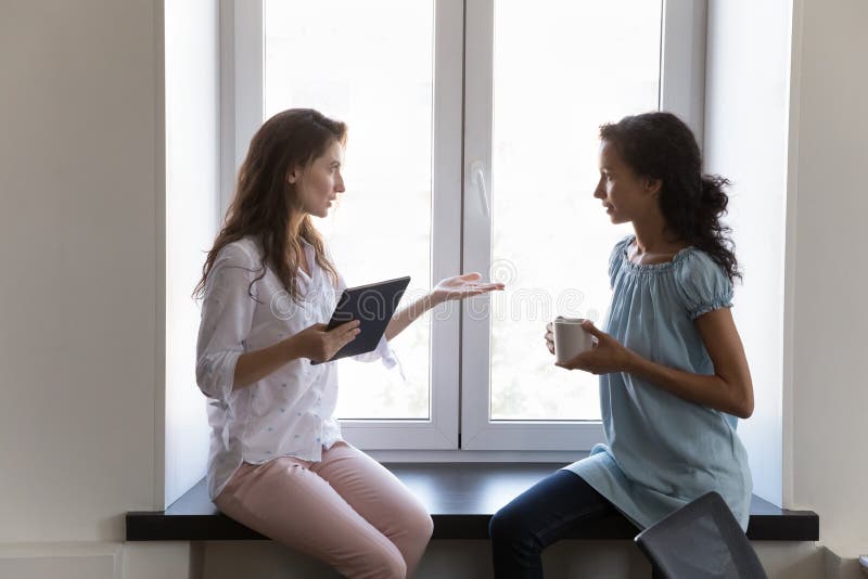 Two diverse coworkers women sitting at office window