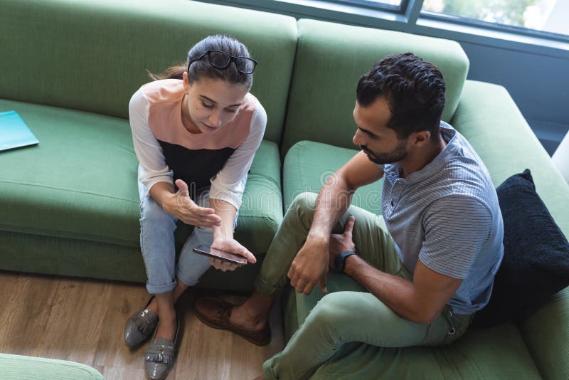Two diverse creative colleagues sitting on sofa using smartphone royalty free stock images