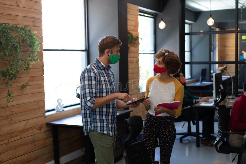Two diverse creative colleagues wearing face masks talking in office royalty free stock photo