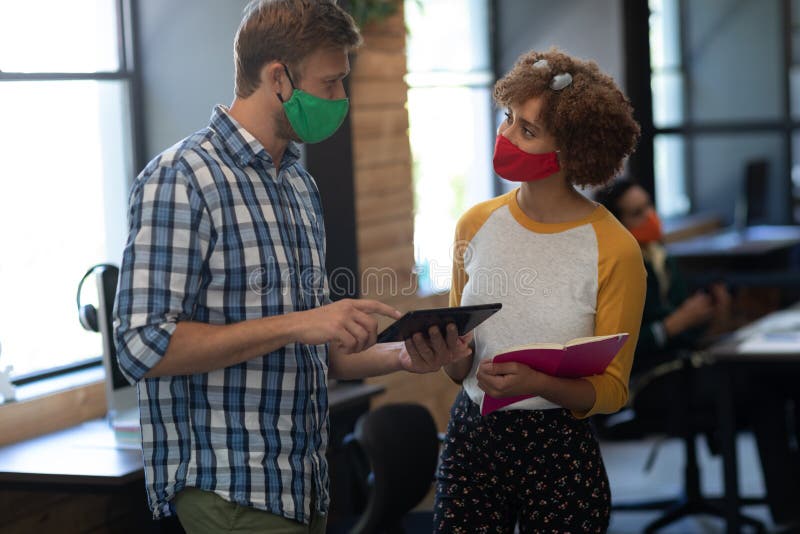 Two diverse creative colleagues wearing face masks talking in office royalty free stock image