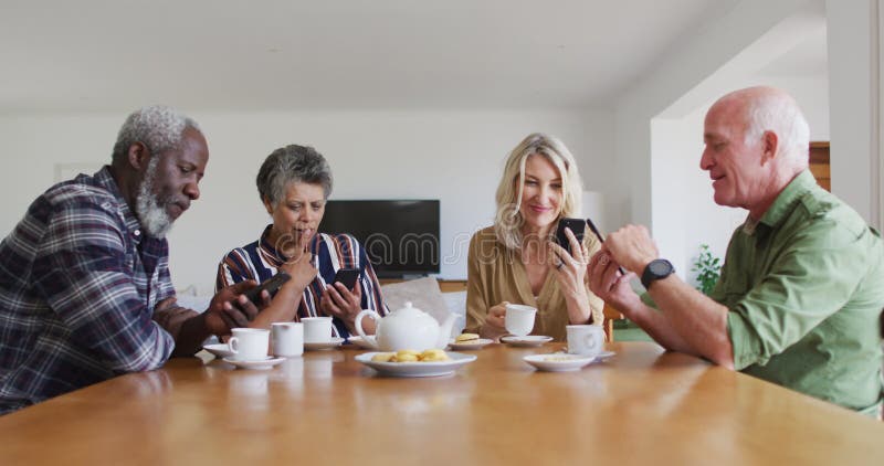 Two diverse senior couples sitting by a table drinking tea using smartphones at home