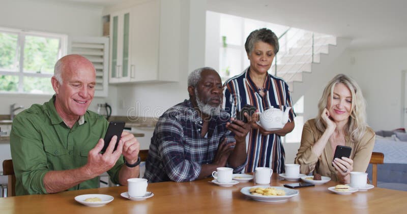 Two diverse senior couples sitting by a table drinking tea using smartphones at home