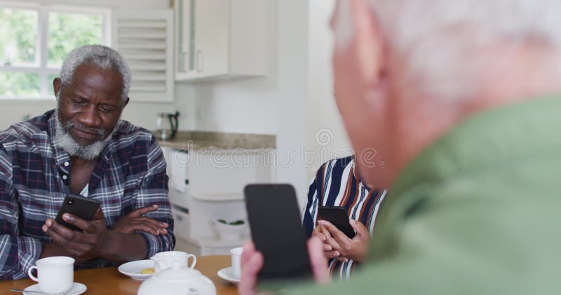 Two diverse senior couples sitting by a table drinking tea using smartphones at home