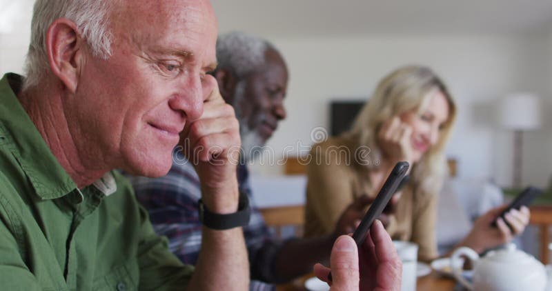 Two diverse senior couples sitting by a table drinking tea using smartphones at home