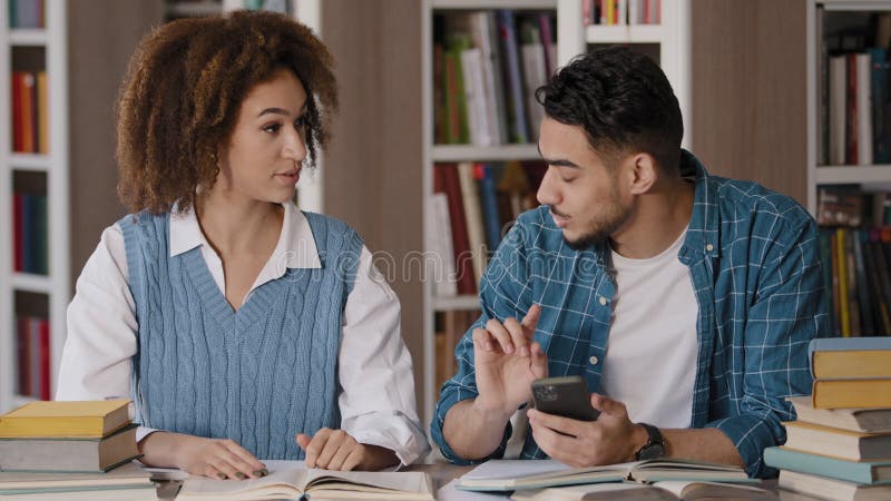 Two diverse students sitting in library preparing for exam young guy enthusiastically plays on phone distracted from stock video