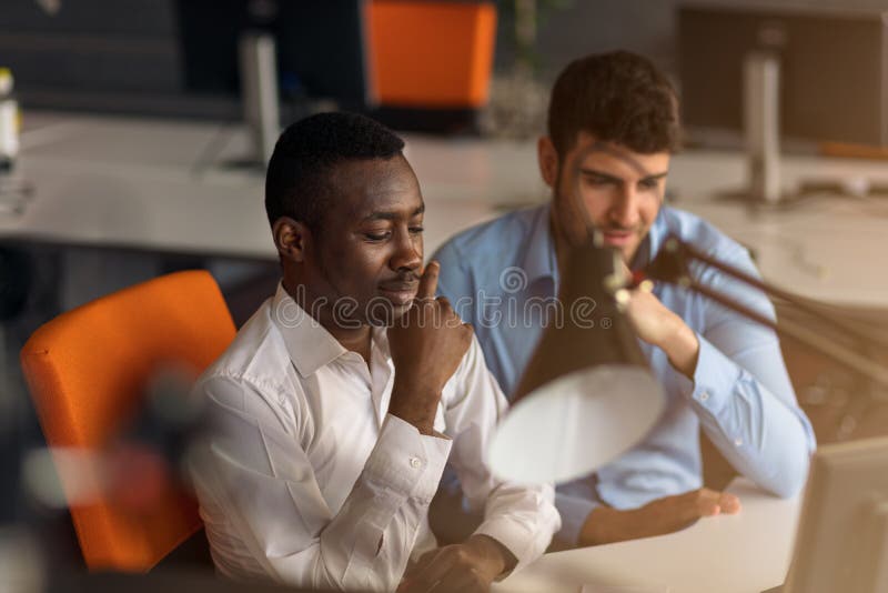 Two diverse work colleagues smiling while sitting together in an office discussing paperwork and working on a laptop stock photo