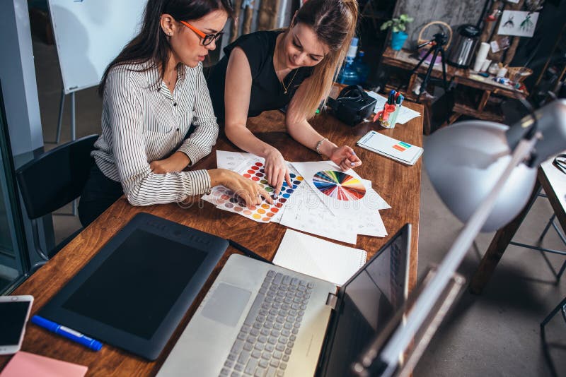 Two female architects working together using color swatches sitting at desk with laptop, graphic tablet in design studio royalty free stock photography