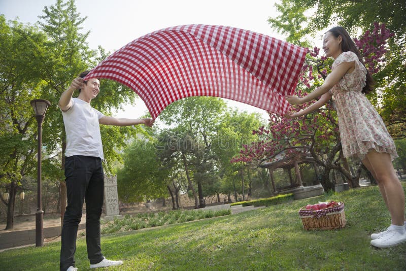 Two friends opening up a checkered blanket and getting ready to have a picnic in the park