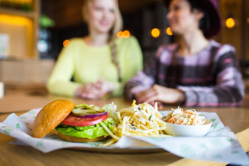 Two girl - eating hamburger and drinking in a fast food diner; focus on the meal stock photo Two girl - eating hamburger and drinking in a fast food diner; focus on the meal stock photo