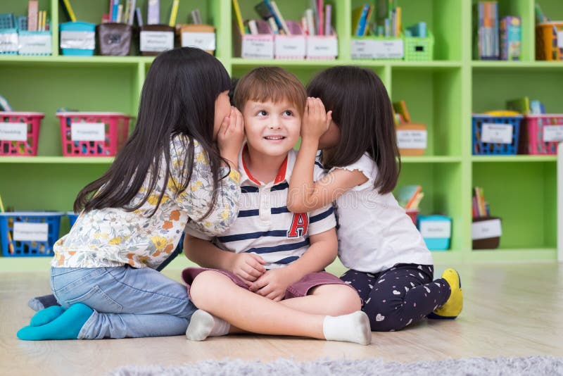 Two girl kids whisper secret at ear of boy in library at kindergarten preschool,Fun and happy children,back to school concept
