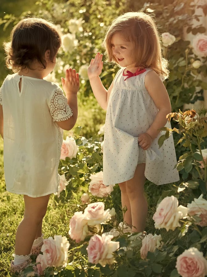 Two girls baby at blossoming rose flowers. Children playing patty cake in summer garden on sunny day. Friends and