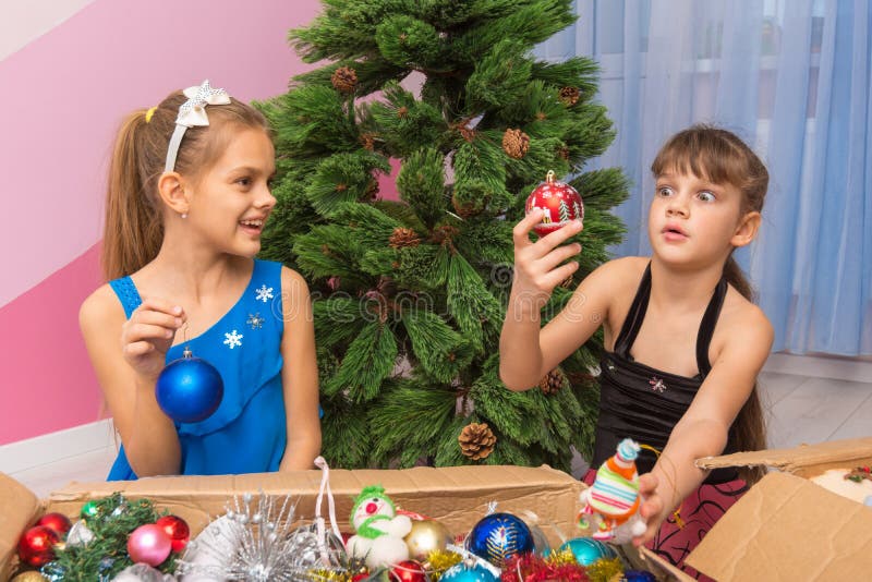 Two girls pull Christmas toys out of the boxes in front of an artificial Christmas tree