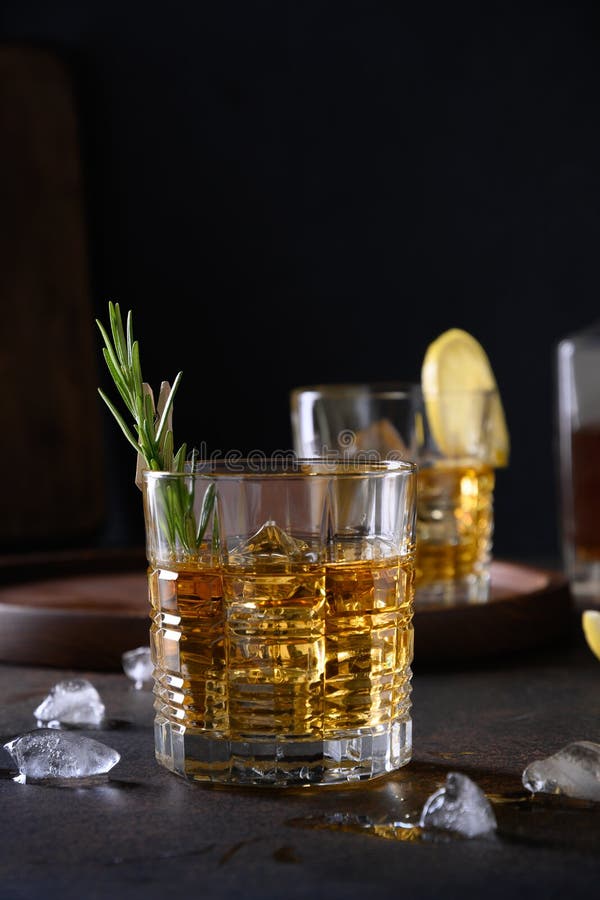 Two glasses of cold whiskey with rosemary, lemon peels on dark brown background. Vertical orientation