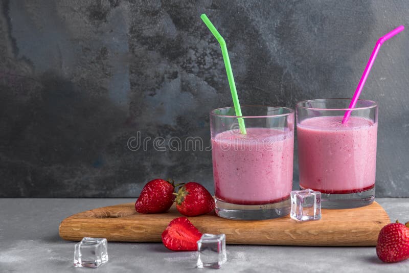 Two glasses of strawberry cocktail with straws on a wooden board and gray background with ice cubes, refreshment drinks for summer
