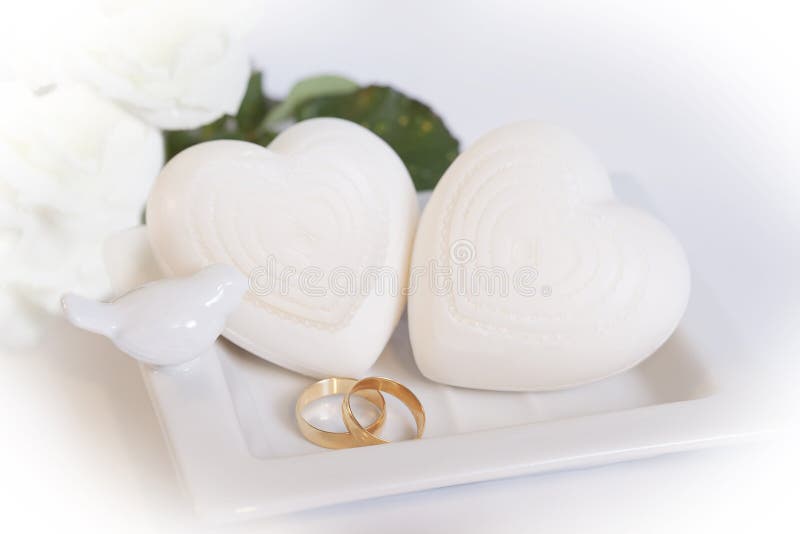 Due bande di nozze d'oro in ceramica bianca vassoio con due a forma di cuore, saponi e piccoli uccelli, e due rose bianche sfocato sullo sfondo.