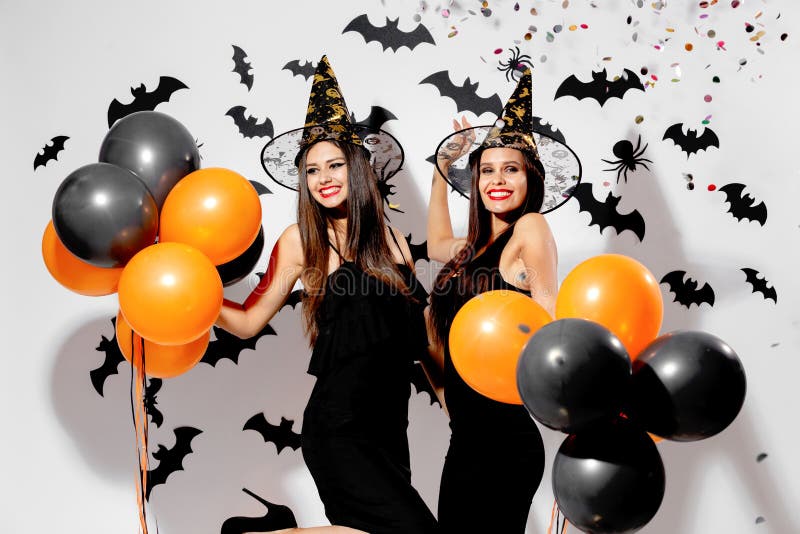 Two gorgeous women in witches hats smile and hold black and orange balloons on a Halloween background. Confetti around .