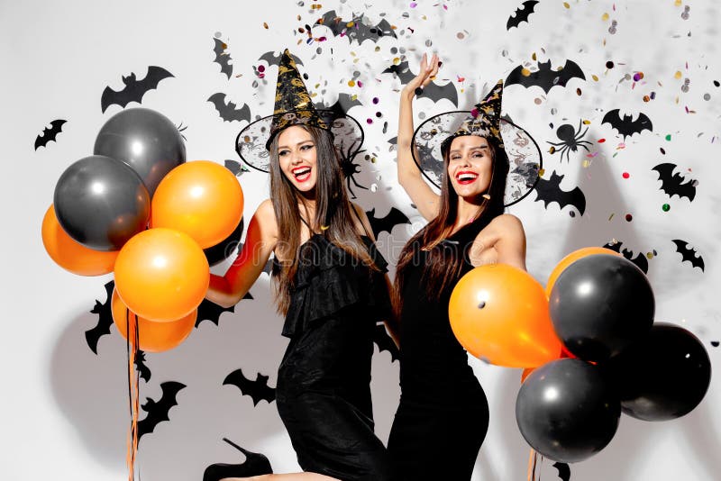 Two gorgeous women in witches hats smile and hold black and orange balloons on a Halloween background. Confetti around .