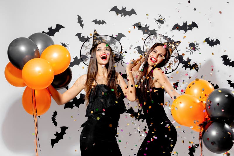 Two gorgeous women in witches hats smile and hold black and orange balloons on a Halloween background. Confetti around .