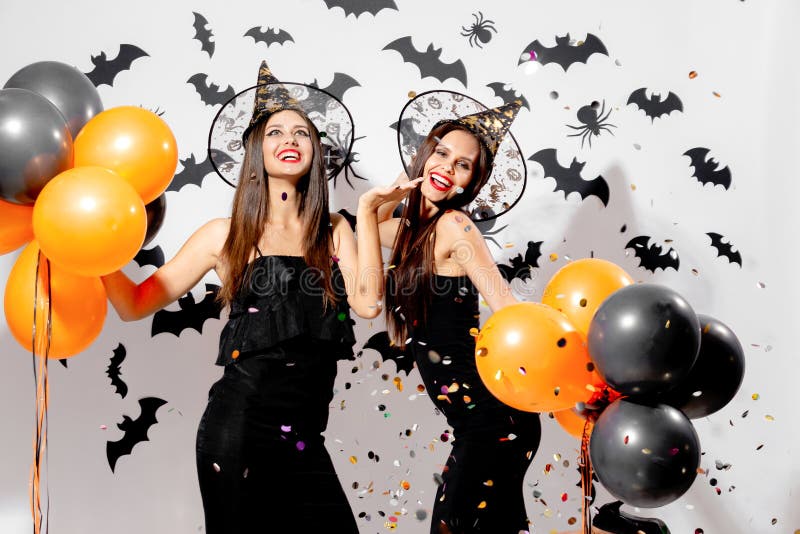 Two gorgeous women in witches hats smile and hold black and orange balloons on a Halloween background. Confetti around .