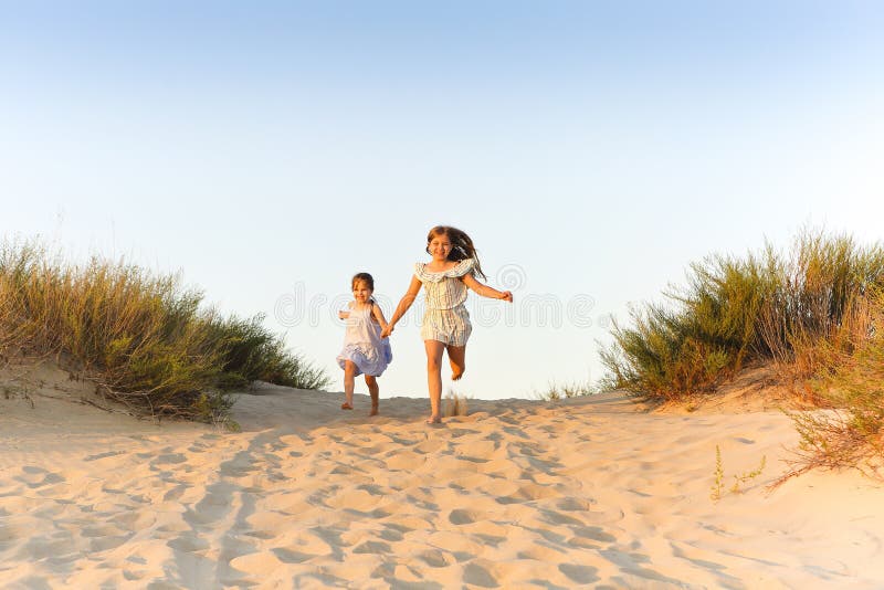 Two happy cheerful little sisters running hand in hand on sand on sunny evening beach