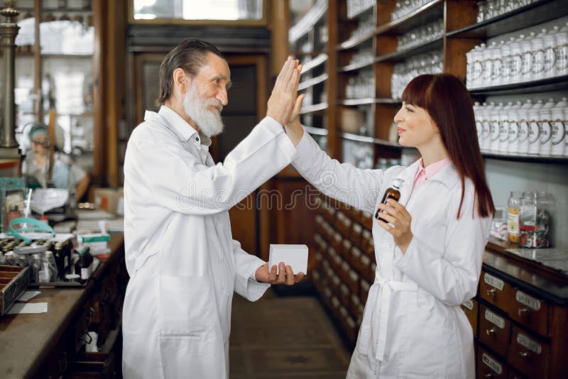 Two happy colleages pharmacists, senior man and young woman, working in old ancient drugstore, smiling and giving high