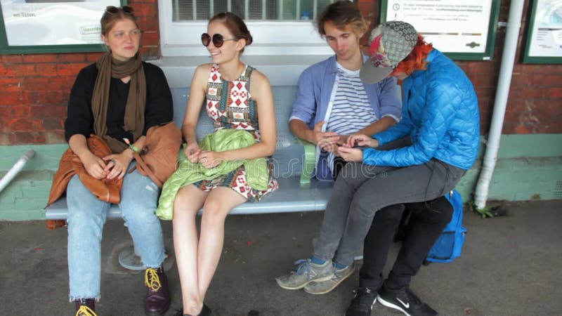 Two happy hipster young girls and a young couple waiting a train on the railway station. They are siting, talking. Exchanging opinions, joking and fooling