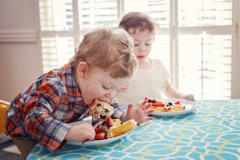 Two happy kids twins boy girl eating breakfast waffles with fruits sitting at table