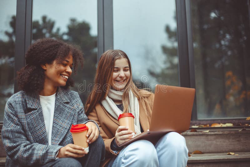 Two happy multiracial teen girls looking at laptop and laughing while drinking coffee in autumn park