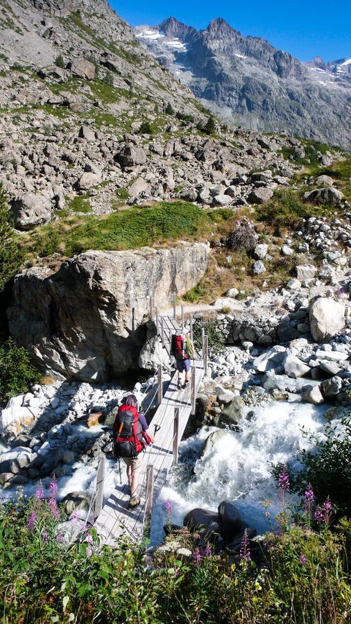 Two hikers and mountain climbers cross a small stream over an old wooden bridge on their way to a base camp in the French Alps. Two hikers and mountain climbers cross a small stream over an old wooden bridge on their way to a base camp in the French Alps