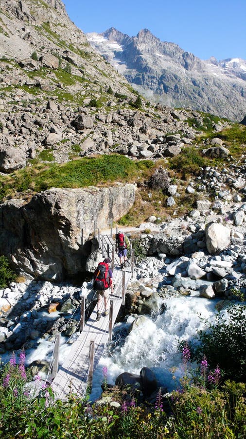 Two hikers and mountain climbers cross a small stream over an old wooden bridge on their way to a base camp in the French Alps. Two hikers and mountain climbers cross a small stream over an old wooden bridge on their way to a base camp in the French Alps