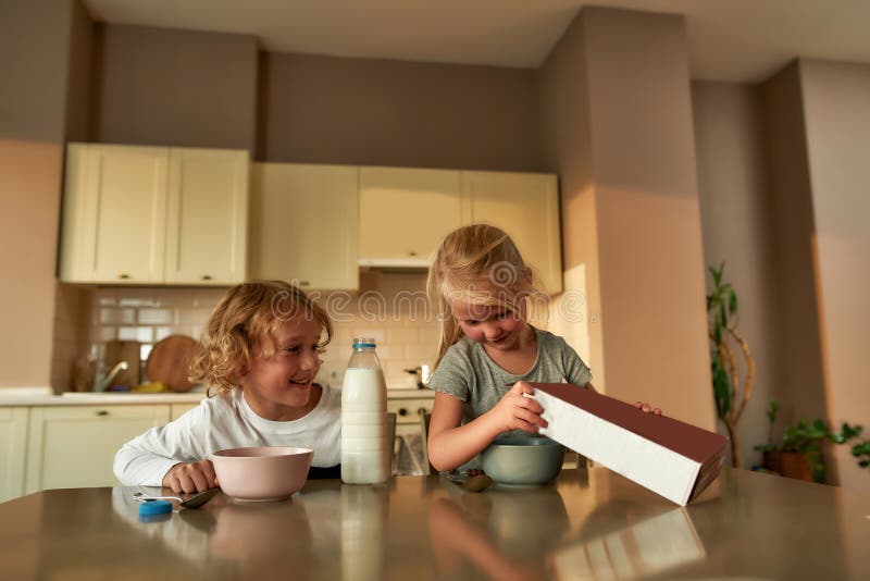 Two joyful little kids pouring chocolate flakes into a bowl while preparing cereal with milk for breakfast, sitting