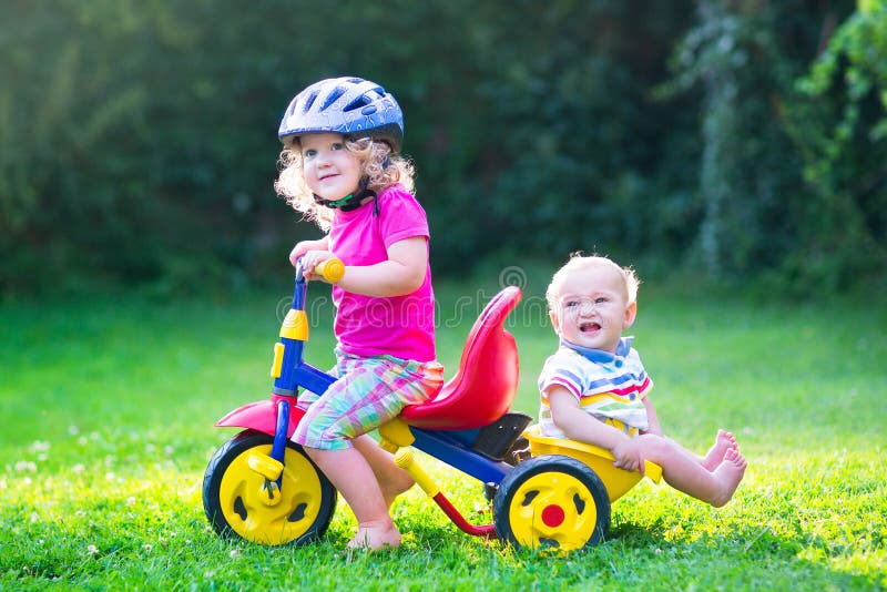 Two happy kids, adorable curly toddler girl and a funny cute baby boy, brother and sister, playing together riding a bike, first colorful tricycle, having fun in the garden on a sunny summer day. Two happy kids, adorable curly toddler girl and a funny cute baby boy, brother and sister, playing together riding a bike, first colorful tricycle, having fun in the garden on a sunny summer day