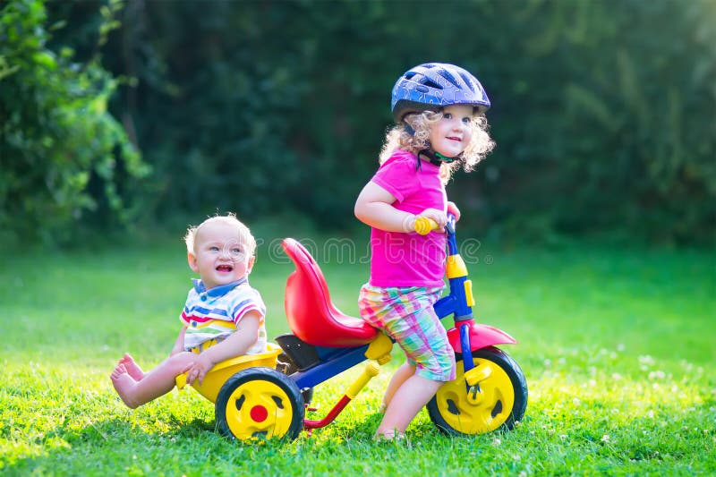 Two happy kids, adorable curly toddler girl and a funny cute baby boy, brother and sister, playing together riding a bike, first colorful tricycle, having fun in the garden on a sunny summer day. Two happy kids, adorable curly toddler girl and a funny cute baby boy, brother and sister, playing together riding a bike, first colorful tricycle, having fun in the garden on a sunny summer day