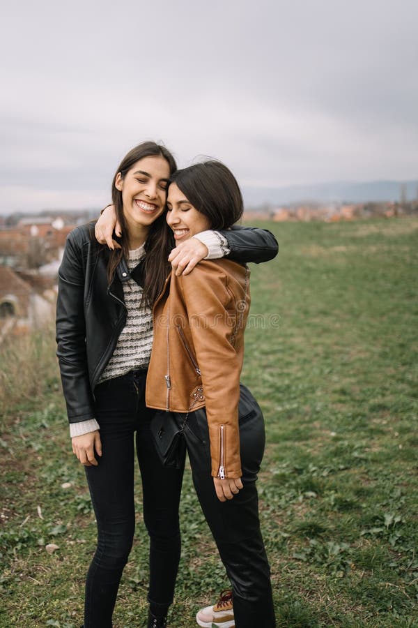 Two laughing girls hugging on field with city view. Sisters meeting and hugging on a field during spring season.