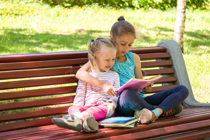 Two little girls are sitting on a bench in summer park and reading a book
