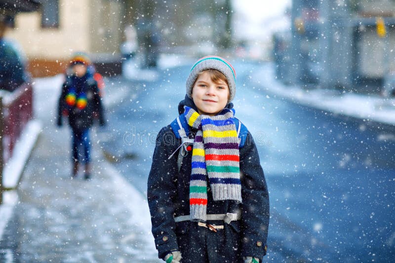 Two little kids boys of elementary class walking to school during snowfall. Happy children having fun and playing with first snow. Siblings ans friends with backpack in colorful winter clothes