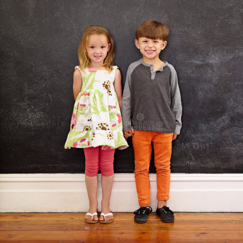 Full length shot of two little kids standing together against a blackboard. Little boy and little girl at home looking at camera smiling. Full length shot of two little kids standing together against a blackboard. Little boy and little girl at home looking at camera smiling.