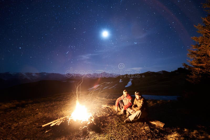 Two male tourists have a rest in his camp near the forest at night. Guys sitting near campfire and tent under beautiful night sky full of stars and the moon, and enjoying night scene in the mountains. Two male tourists have a rest in his camp near the forest at night. Guys sitting near campfire and tent under beautiful night sky full of stars and the moon, and enjoying night scene in the mountains