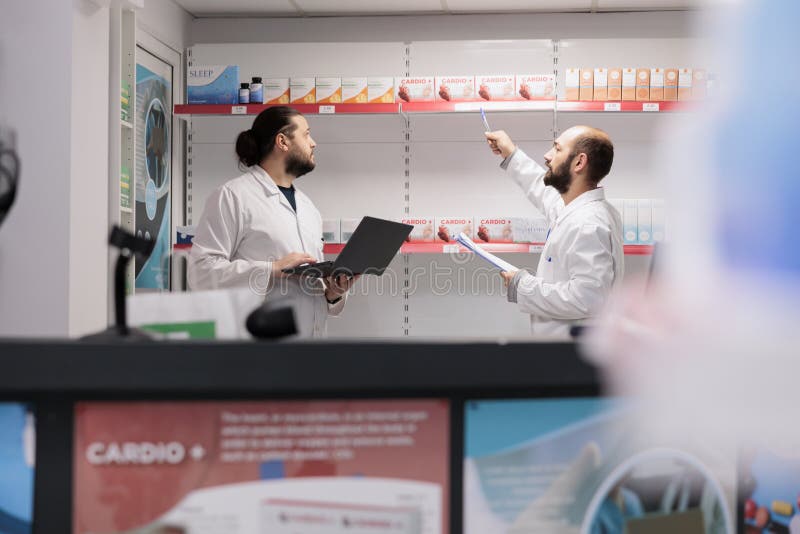 Two men pharmacists doing medicaments inventory checking drugs packages, typing vitamins information on laptop. Pharmacy is a reliable and convenient location for filling prescription medication