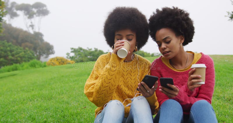 Two mixed race women drinking coffee and listening music in park