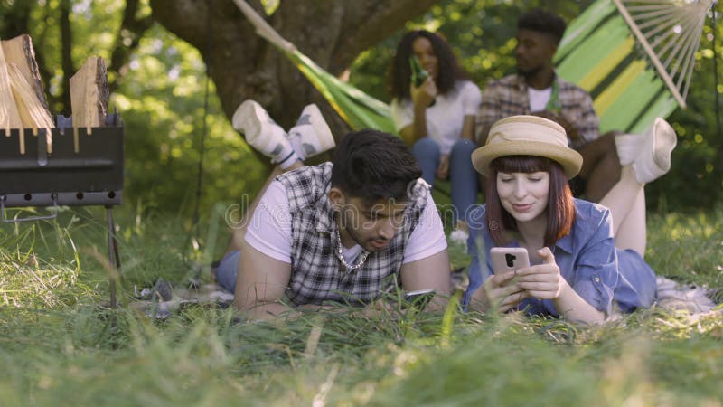 Two multiracial couples relaxing, chatting and drinking on picnic outdoors