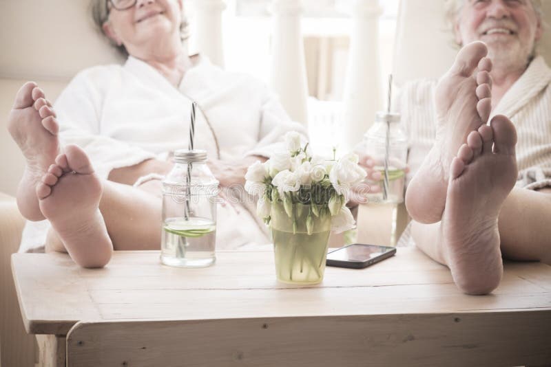 two pensiones or seniors sitted indoor in a resort or hotel doing a beauty treatment - close up of feet on the table with cocktail.