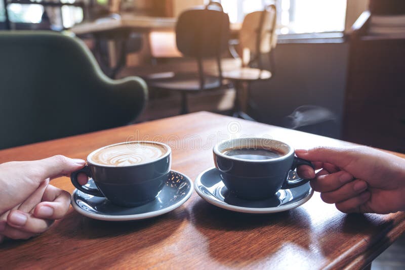 Two people`s hands holding coffee and hot chocolate cups on wooden table in cafe