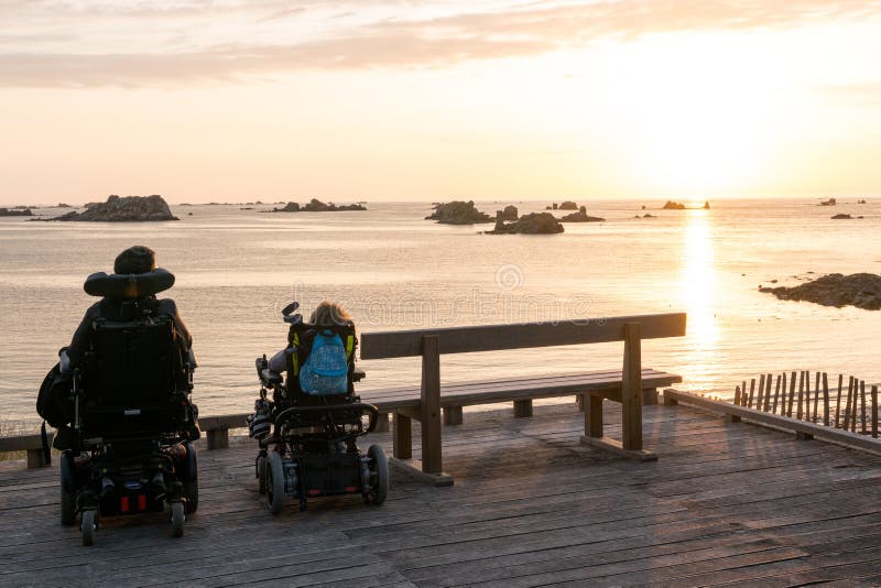 Two people in wheelchairs enjoy the opportunity to watch a sunset over the beach and ocean