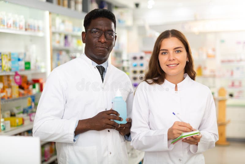 Pharmacists in white gowns standing in salesroom of drugstore and smiling. Pharmacists in white gowns standing in salesroom of drugstore and smiling