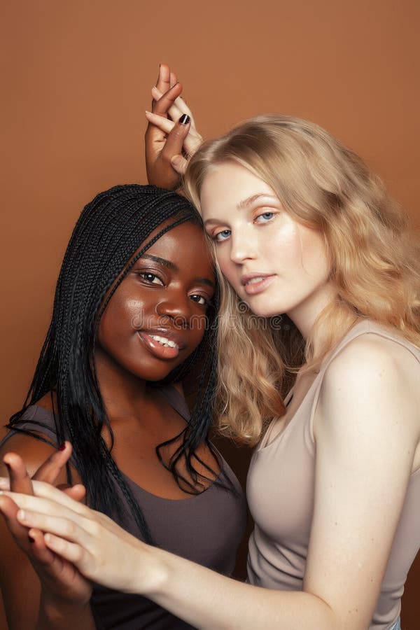 Two pretty girls african and caucasian blond posing cheerful together on brown background, ethnicity diverse lifestyle